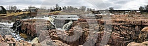 The Big Sioux River flows over rocks in Sioux Falls South Dakota with views of wildlife, ruins, park paths, train track bridge, tr