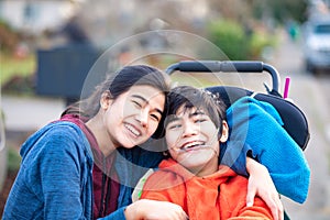 Big sister hugging disabled brother in wheelchair outdoors, smiling photo