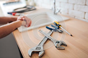 Big and small wrenches on table in kitchen. Screwdriver and nippers. Man`s hands on sink. Daylight.