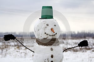 Big smiling snowman with bucket hat, scarf and gloves on white snowy field winter landscape, blurred black trees and blue sky copy