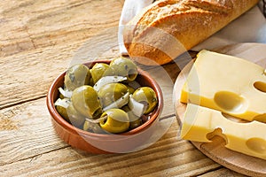 Big Spanish Green Gordal Olives with Herbs and Onions in Earthenware Bowl Baguette Maasdam Cheese on Cutting Board