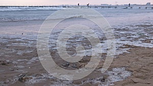 Big stormy waves in the sea. Beautiful seascape with pier, sandy beach and coastal town on background at sunset.