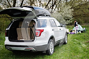 Big SUV car with open trunk and roof rack box against happy young family having fun and enjoying outdoor on picnic blanket at