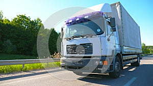 Big truck with cargo trailer driving on highway and transporting goods at sunny day. White lorry riding to destination