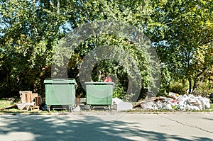 Big two green plastic dumpster garbage cans full of overflow litter polluting the park and street in the city with junk