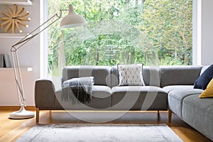 Big white metal lamp standing next to grey corner lounge with blanket and cushion in the real photo bright living room interior wi