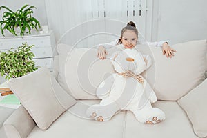 Big white soft bear is on the sofa. Girl is standing behind the sofa, leaning over its back, looking at the camera