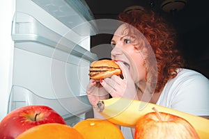 Big woman eat fast food. Red hair fat girl looking inside refrigerator with burger. Unhealthy and healthy food concept with plus