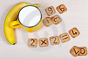 Big yellow cup of milk, banana and funny cookies with numbers on light wooden background. Healthy breakfast for a child.