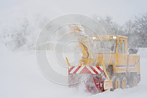 Big yellow snowplough with and helix is cleaning the road while a powerful stream of white snow go out