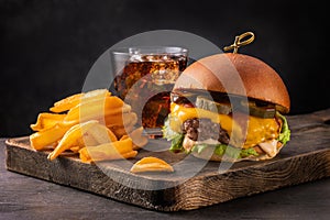 Big yummy burger with cheese, fries and a glass of cola on a wooden board on a black background, angle view.