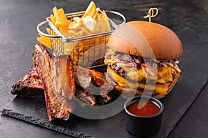 Big yummy burger with double cutlet, fries and fried ribs with barbecue sauce on a black slate background.