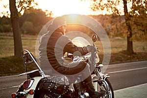 Biker in black leather outfit driving modern powerful motorcycle along sunny road on summer day.