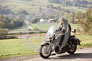 Biker in black leather outfit driving modern powerful motorcycle along sunny road on summer day.