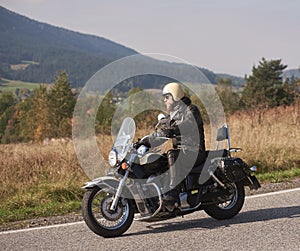 Biker in black leather outfit driving modern powerful motorcycle along sunny road on summer day.