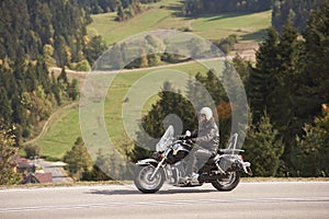 Biker in black leather outfit driving modern powerful motorcycle along sunny road on summer day.