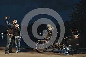 Biker couple made a selfie on smartphone. Stands by a tourer motorcycle with bags. Tourism and vacation. Summer night. Bled lake,