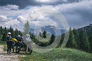 Biker lady with adventure touring motorcycle in full equipment on side of the road, Mountains and forest with rainy clouds on