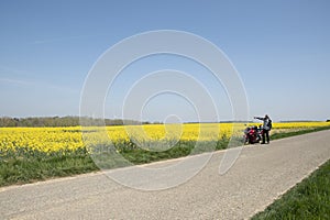 biker with red motorcycle stands next of a blooming yellow field of rapeseed