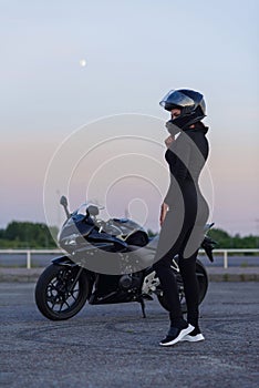 Biker woman in black leather jacket and full face helmet sits near stylish sports motorcycle at urban parking. Traveling