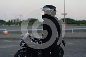 Biker woman in black leather jacket and full face helmet sits near stylish sports motorcycle at urban parking. Traveling