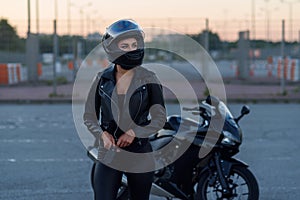 Biker woman in black leather jacket and full face helmet stands near stylish sports motorcycle. Urban parking, sunset in