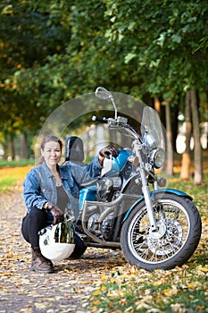 Biker woman with white helmet and jeans outfit portrait, sitting close to motorbike, autumn park
