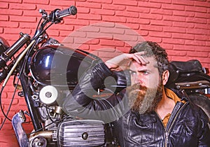 Bikers lifestyle concept. Man with beard, biker in leather jacket near motor bike in garage, brick wall background