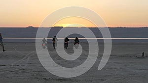 Bikers ride into sunset on lake, river, sea, bay. Motorcyclists passing desert.