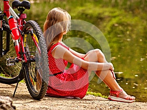 Bikes cycling girl into park. Girl sits leaning on bicycle on shore.