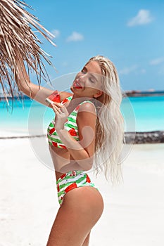 Bikini Model eating watermelon on tropical beach. Pretty slim girl posing on exotic island in turquoise ocean. Blonde in swimwear