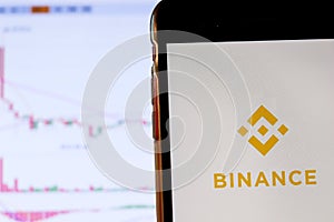 Binance cryptocurrency exchange logo and icon on Phone screen over a laptop display with bitcoin chart. Los Angeles, California