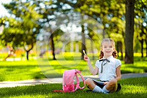 Bingo. Little school girl with pink backpack sitting on grass after lessons and thinking ideas, read book and study