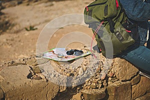 Binoculars, compass on map near backpack of a tourist sitting on the cliff, enjoying his hiking on the adventure travel.