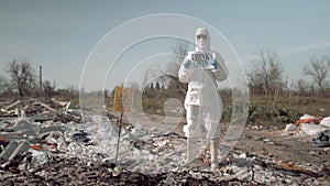 Biological hazard, young woman in uniform and protective glasses holding poster think green standing at trash dump in