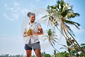 Bipoc man enjoys fresh coconut on sunny tropical beach. Male quenches thirst with natural drink, relaxes by palm trees