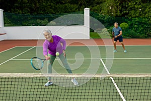 Biracial active senior woman with senior man playing tennis in tennis court against trees