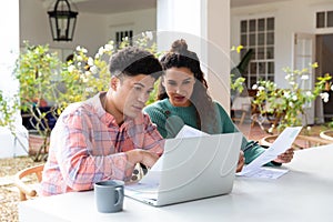 Biracial couple sitting on garden terrace outside house using laptop and paying bills