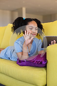 Biracial elementary girl waving over smart phone during video call while lying on couch in school