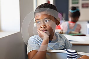 Biracial elementary schoolboy with hand on chin looking away while sitting at desk in classroom