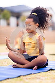 Biracial elementary schoolgirl meditating while sitting on exercising mat at school ground