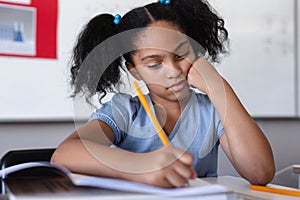 Biracial elementary schoolgirl writing on book at desk in classroom