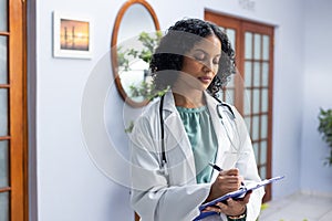 Biracial female doctor wearing stethoscope holding clipboard and taking notes at doctor's office