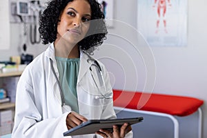 Biracial female doctor wearing sthethoscope, using tablet at doctor's office