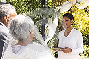Biracial female marriage officiant using tablet with senior couple during wedding in sunny garden