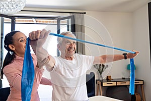 Biracial female physiotherapist assisting caucasian senior man stretching resistance band at home