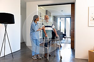 Biracial female physiotherapist assisting caucasian senior man in walking with walker at home