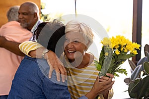 Biracial happy senior guests with bouquet embracing friends at entrance in nursing home