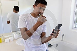 Biracial man brushing teeth and using smartphone in bathroom at home