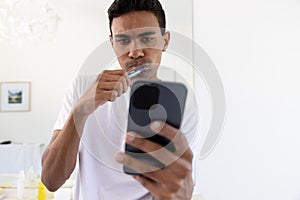 Biracial man brushing teeth and using smartphone in bathroom at home